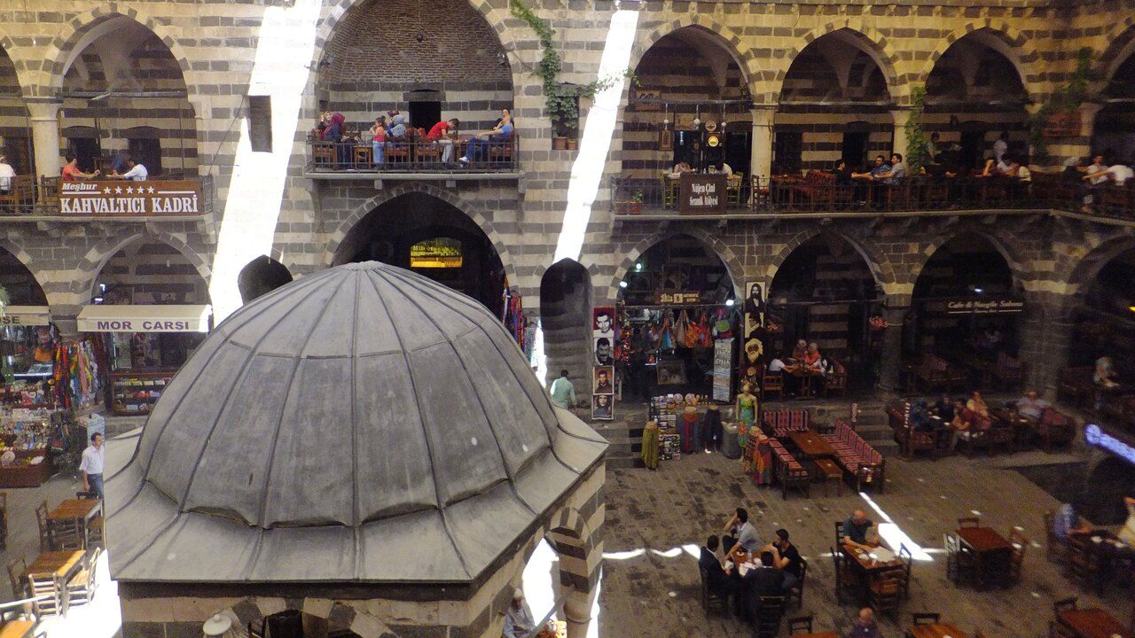 Wide view of Hasan Pasha Han's courtyard in Diyarbakir, with stone arcades, a domed fountain and traditional cafes.