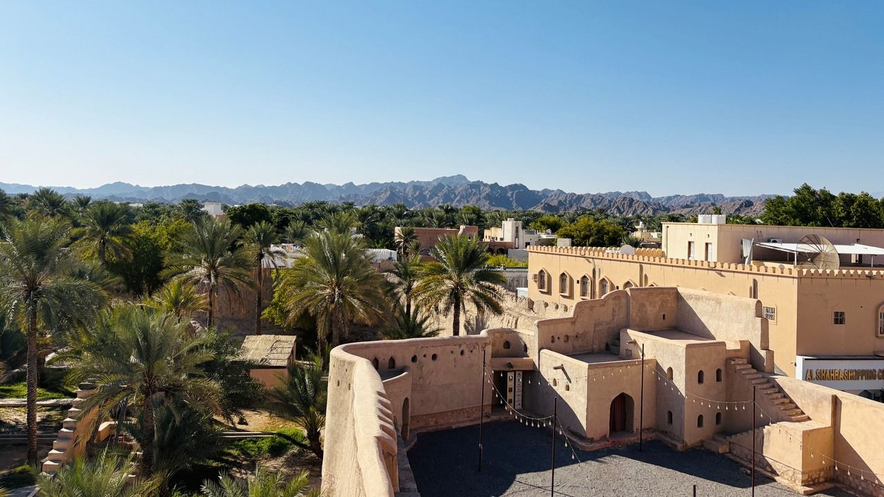 Wide view of Nizwa Fort surrounded by palm trees with the Hajar Mountains in the background, Oman.