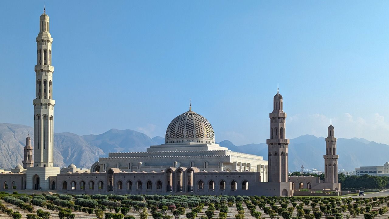 Wide view of Sultan Qaboos Grand Mosque in Muscat, Oman, with its central dome, minarets and Hajar Mountains in the background.