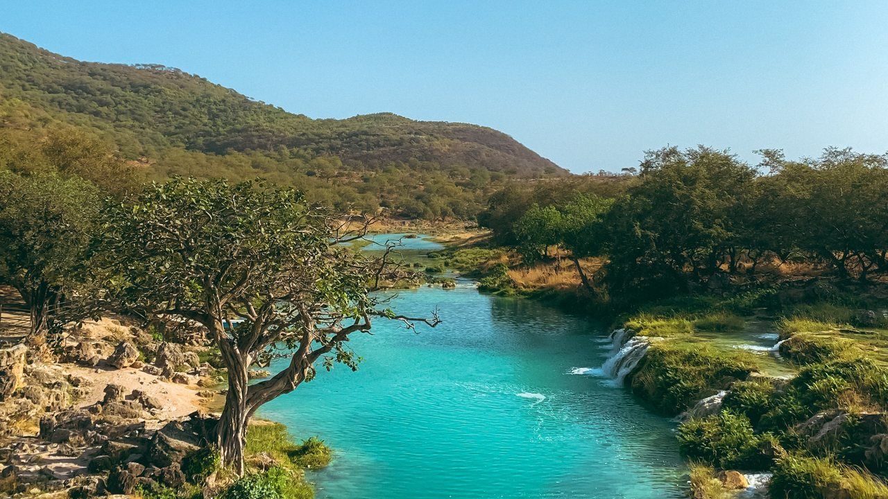 Wide scenic view of turquoise water, small waterfalls and lush mountain scenery at Wadi Darbat near Salalah, Oman.